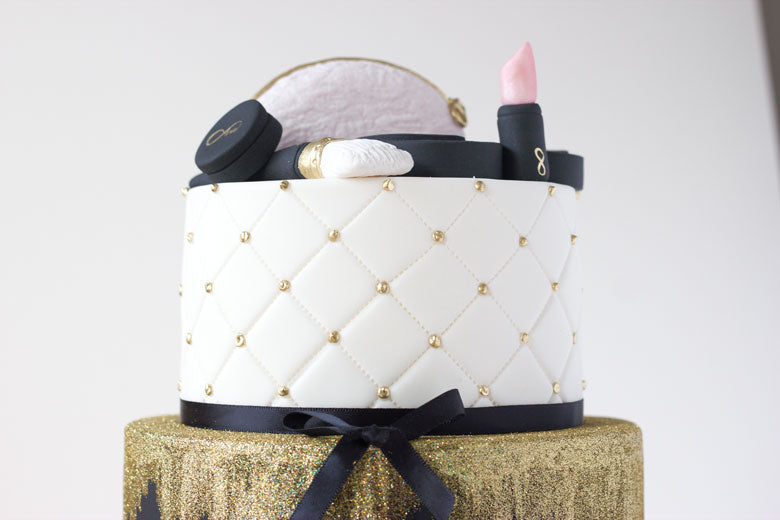 Black and gold makeup cake with gold glitter and quilting decorations. Two tiered cake with edible fondant lipstick, eye shadow palette, mirror, mini makeup bag, makeup brush and nail polish
