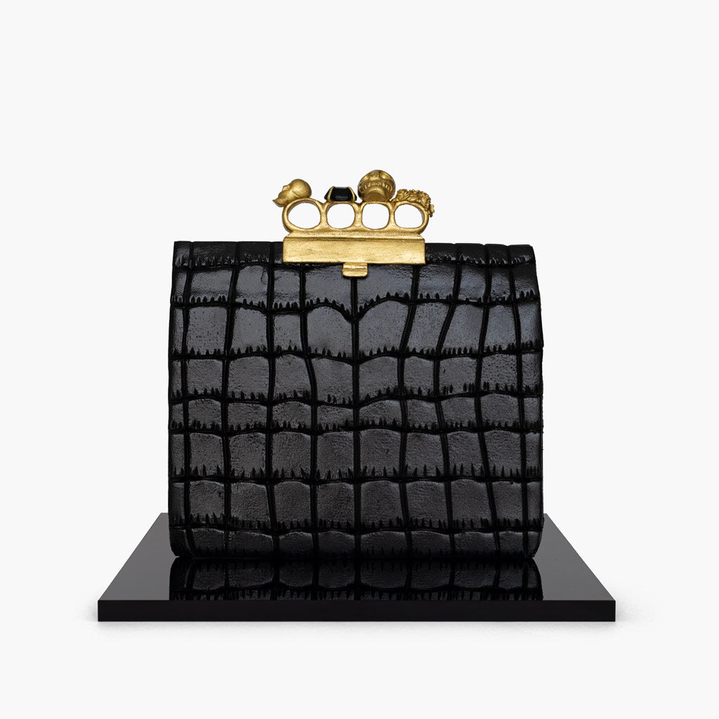 Alexander McQueen knuckle clutch handbag cake. The handbag cake is enveloped in a black fondant leather-like shiny finish. Finished with gold knuckle rings on top.