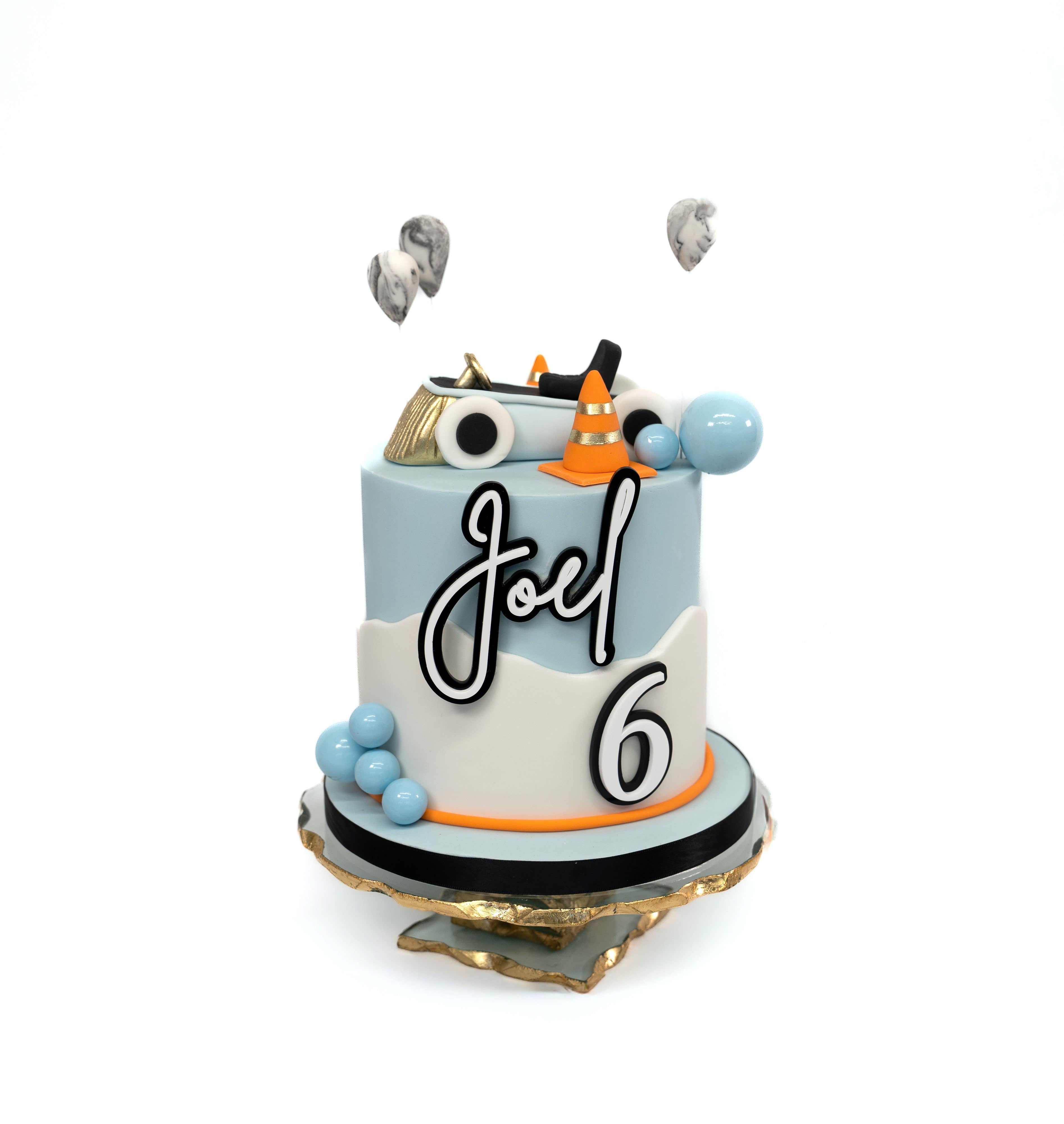 A pastel blue and white car-themed birthday cake featuring an edible car topper, balloons, traffic cones and a personalised name and age design.