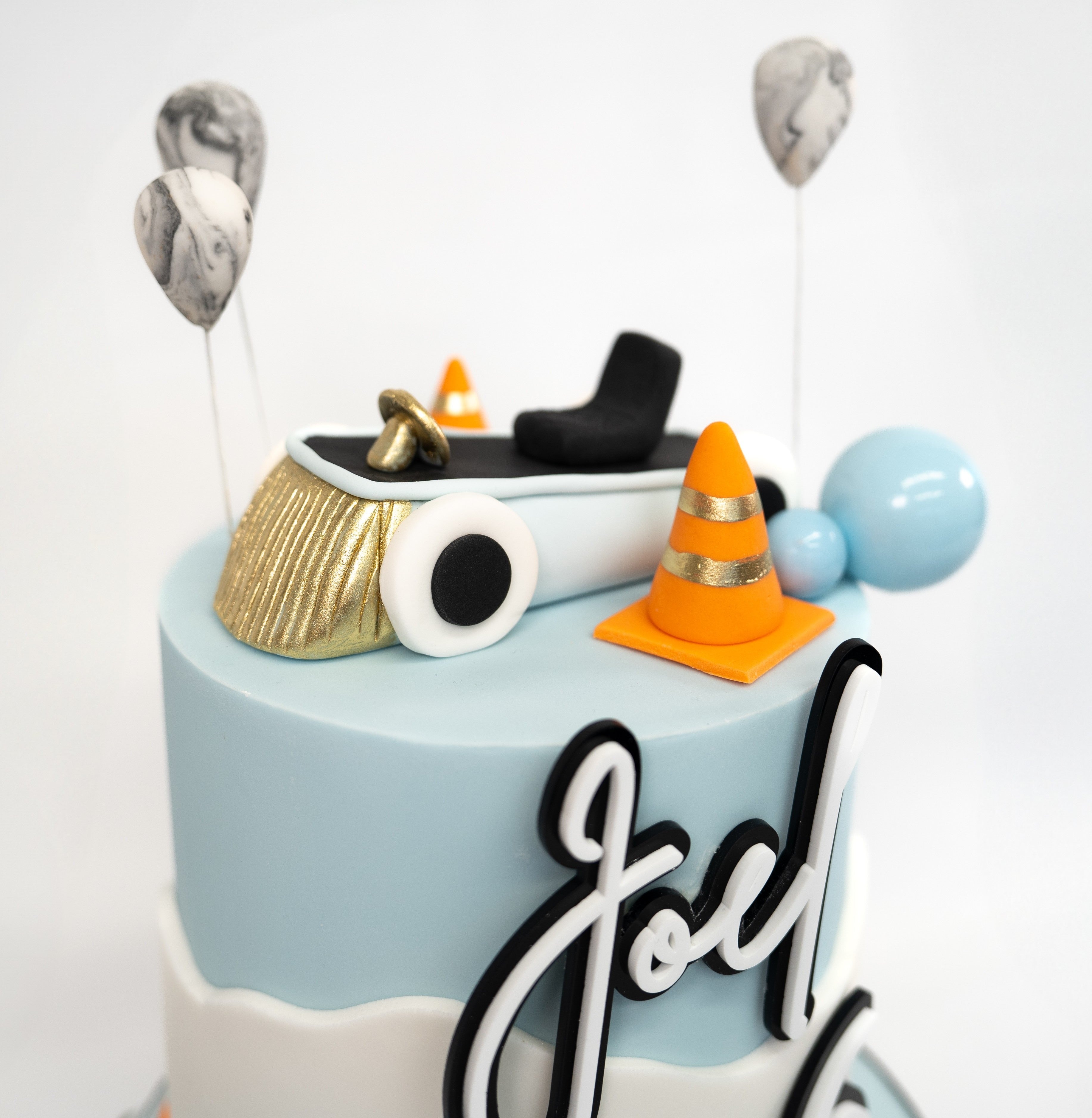 A pastel blue and white car-themed birthday cake featuring an edible car topper, balloons, traffic cones and a personalised name and age design.