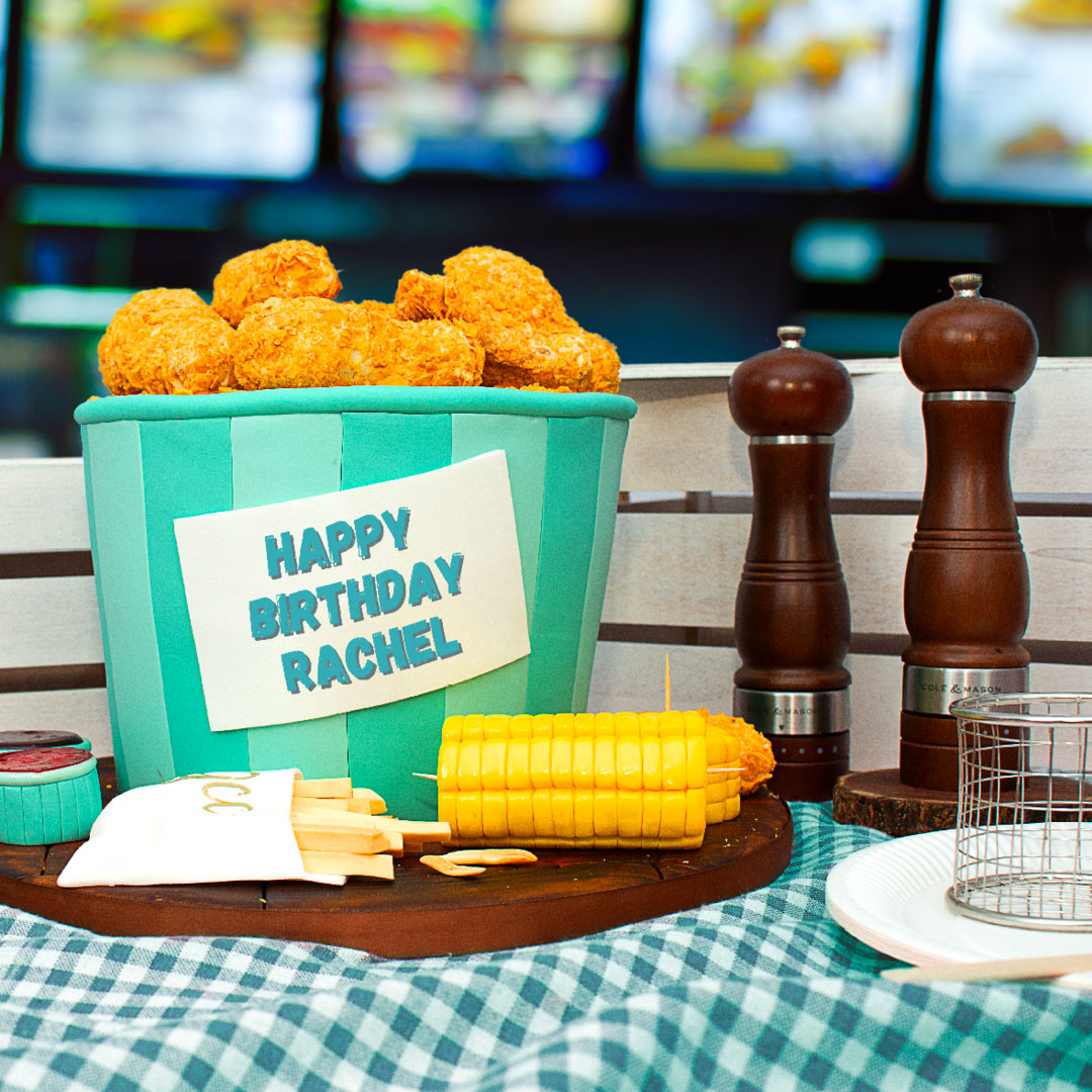 Fried chicken-themed birthday cake featuring a bucket filled with edible chicken pieces, fries, corn on the cob and dipping sauces