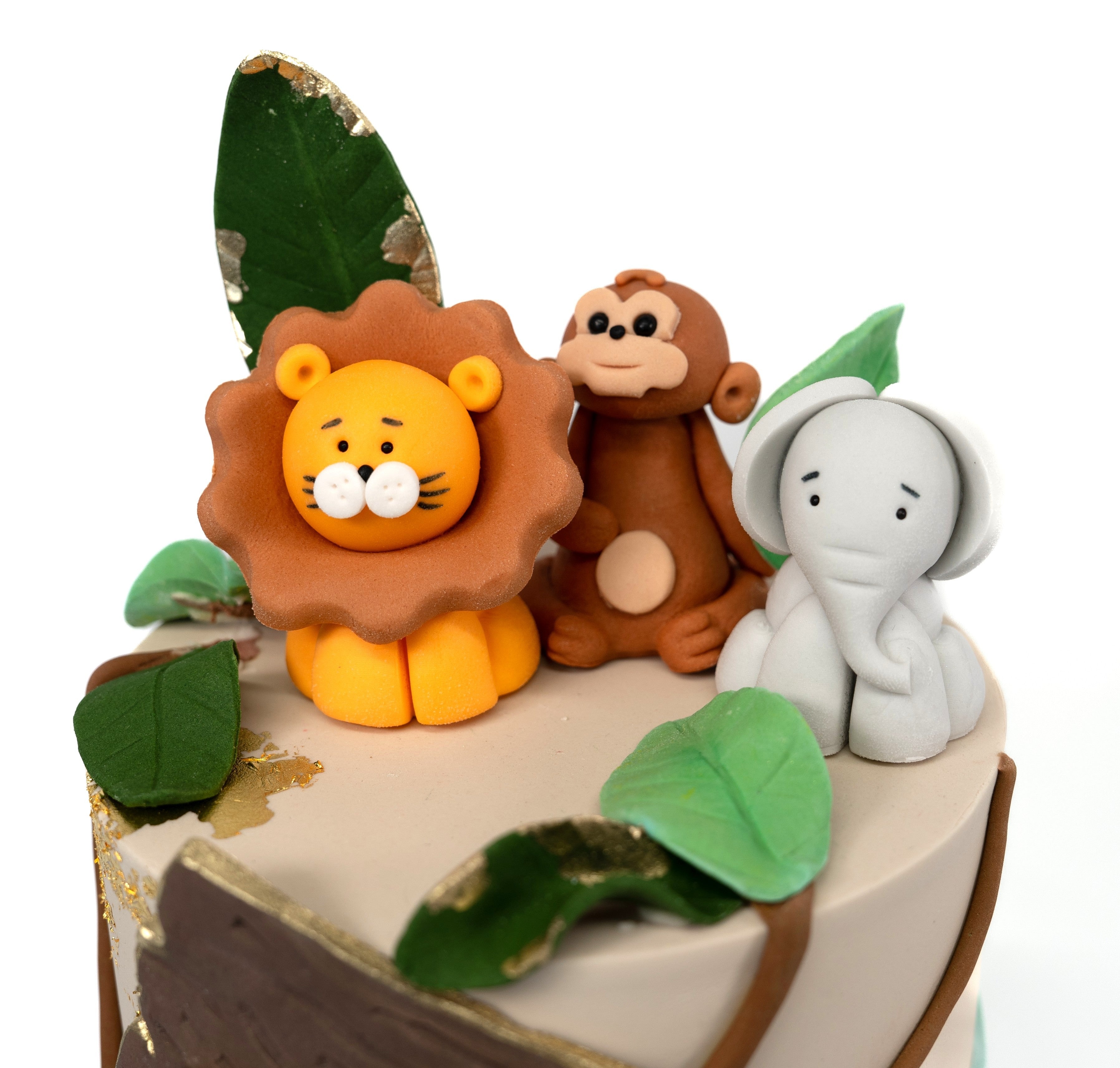 A whimsical jungle-themed cake decorated with fondant animals (kion monkey, elephant, gifaffe) green leaves, gold leaf accents and a wooden-style personalised name plaque