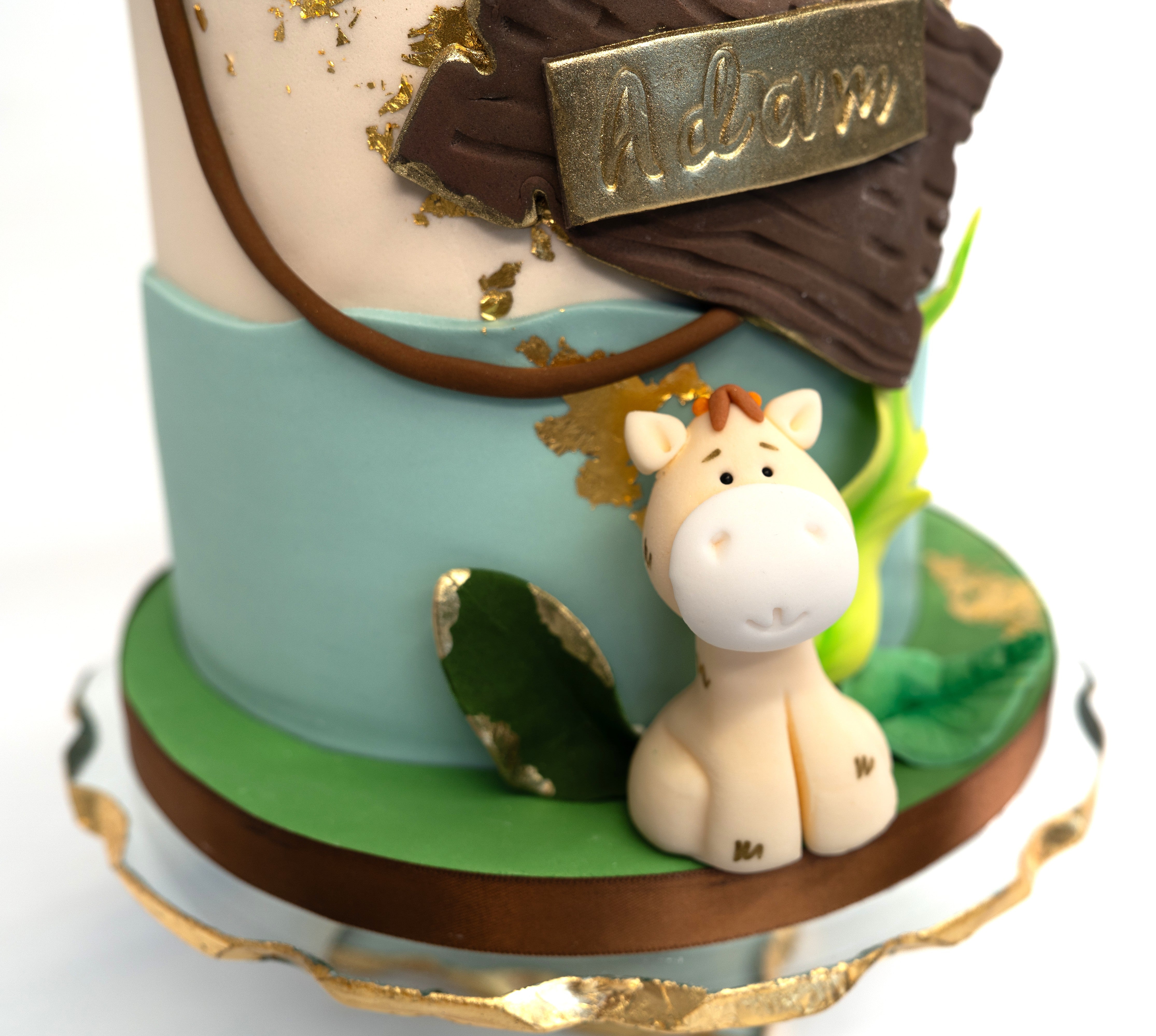 A whimsical jungle-themed cake decorated with fondant animals (kion monkey, elephant, gifaffe) green leaves, gold leaf accents and a wooden-style personalised name plaque