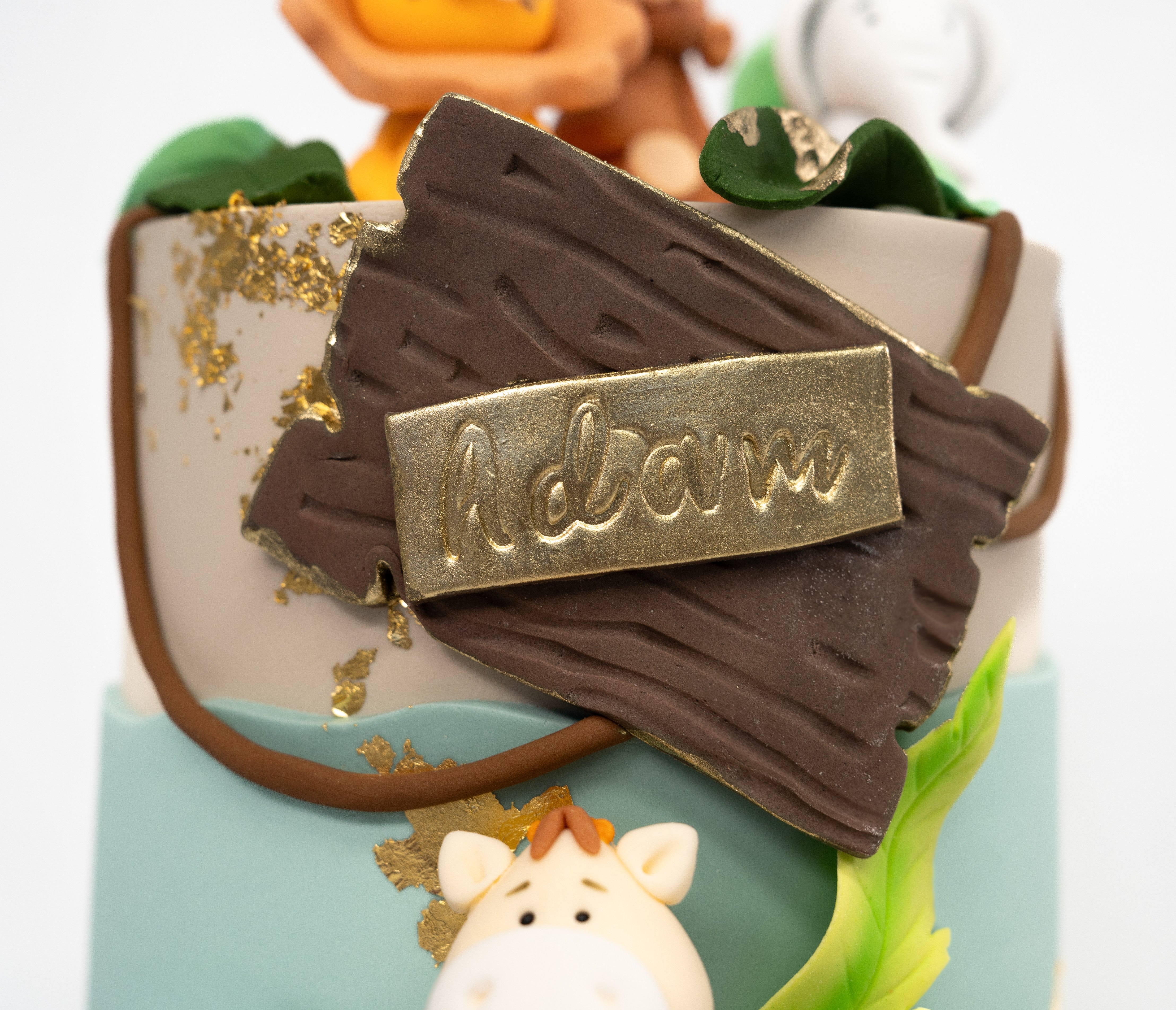 A whimsical jungle-themed cake decorated with fondant animals (kion monkey, elephant, gifaffe) green leaves, gold leaf accents and a wooden-style personalised name plaque