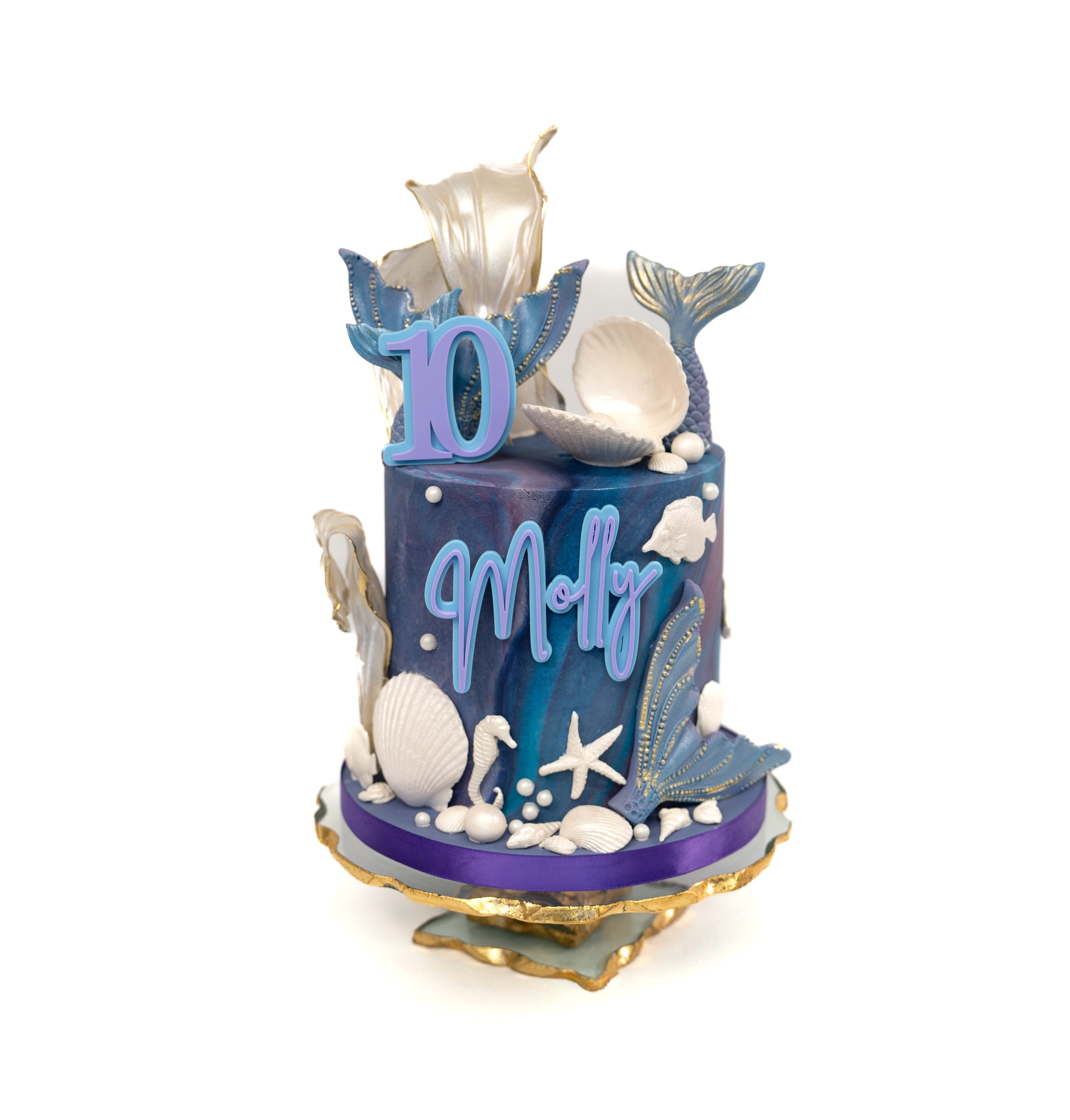 An elegant mermaid-themed birthday cake featuring marbled blue and purple tones, gold-accented mermaid tails, edible seashells, starfish and personalised name and age details