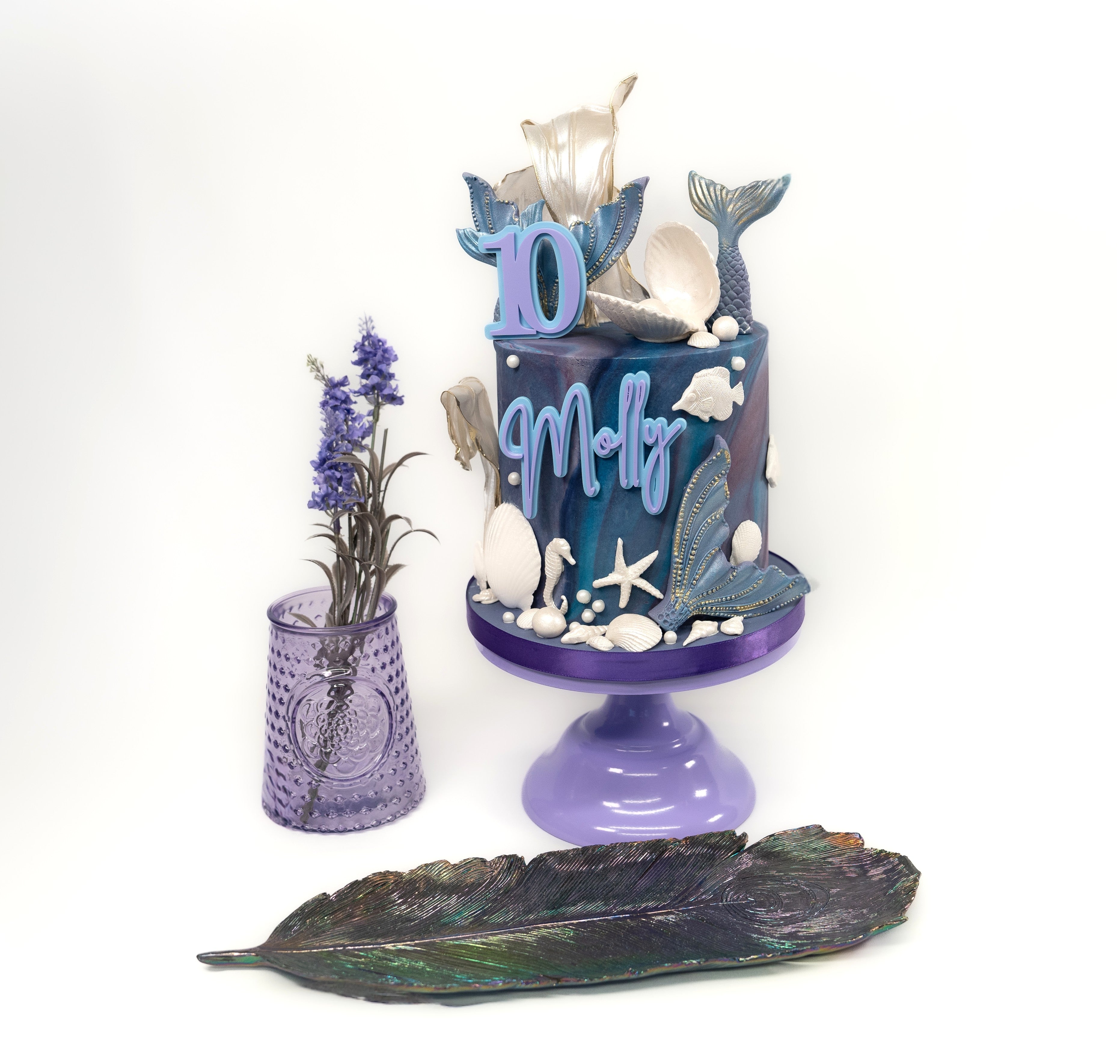 An elegant mermaid-themed birthday cake featuring marbled blue and purple tones, gold-accented mermaid tails, edible seashells, starfish and personalised name and age details