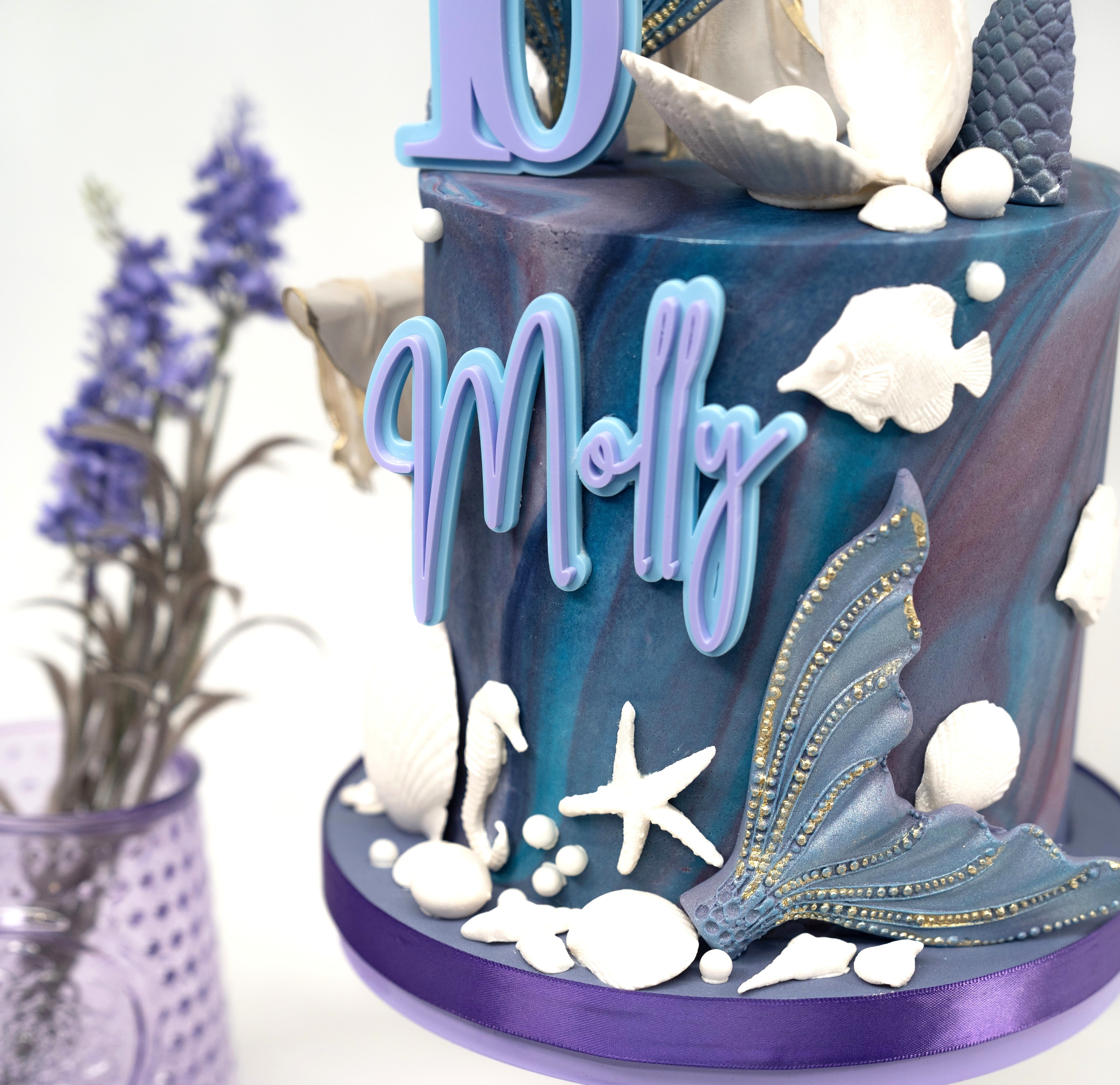 An elegant mermaid-themed birthday cake featuring marbled blue and purple tones, gold-accented mermaid tails, edible seashells, starfish and personalised name and age details