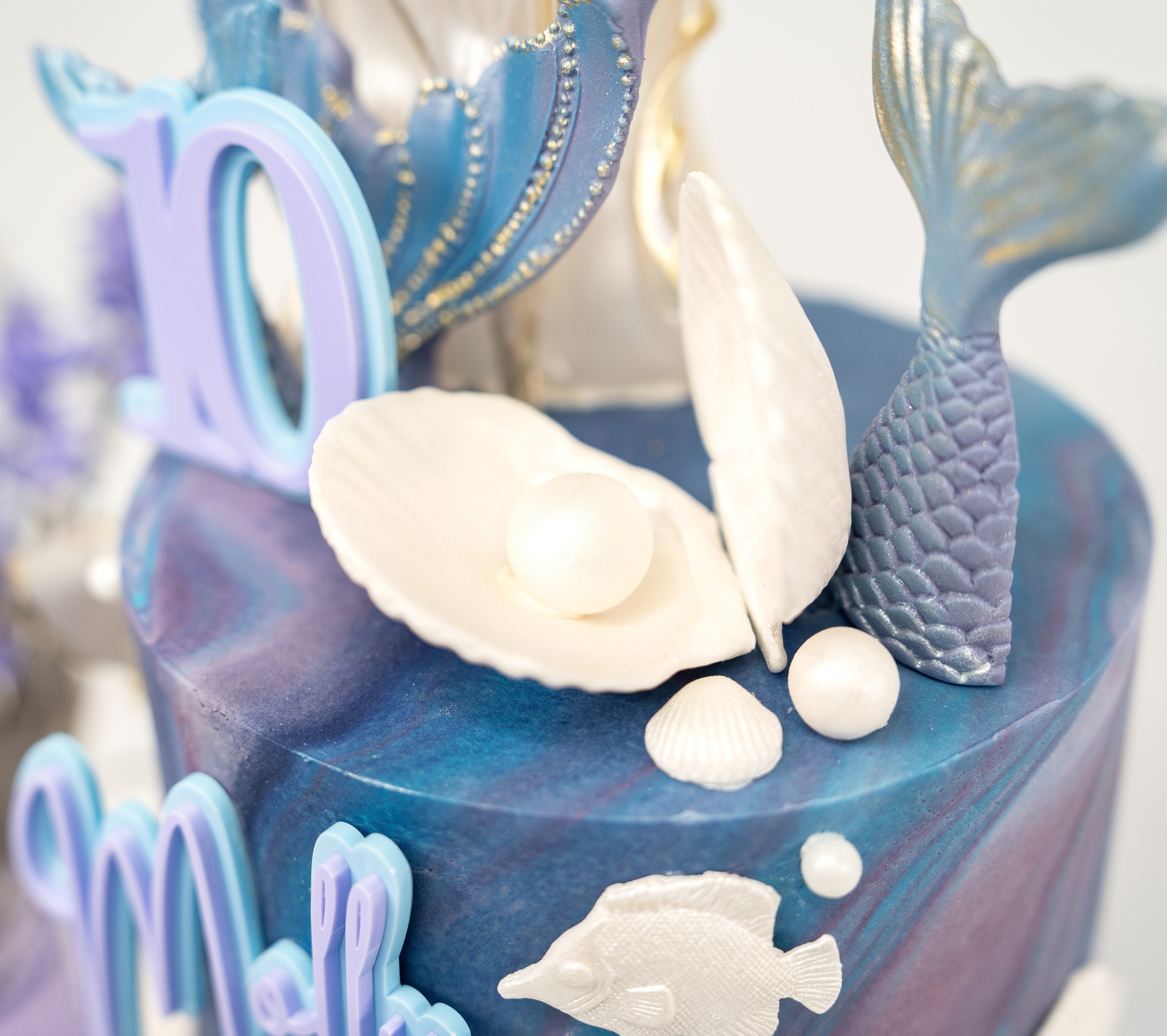 An elegant mermaid-themed birthday cake featuring marbled blue and purple tones, gold-accented mermaid tails, edible seashells, starfish and personalised name and age details