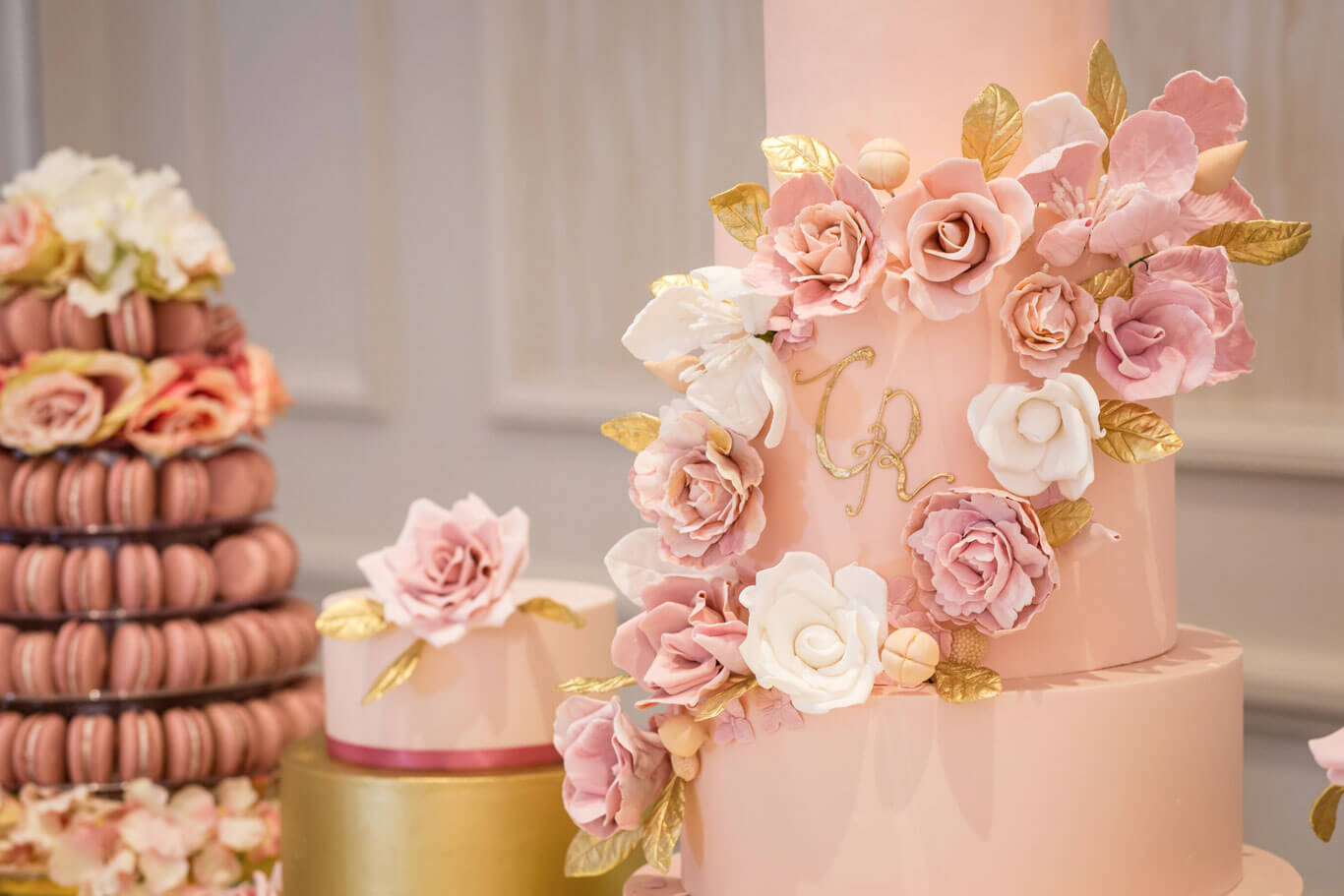 a gordon ramsay wedding cake closeup with initials hand painted in gold surround by elegant handmade sugar flowerss in pink and white as part of a dessert station for a wedding