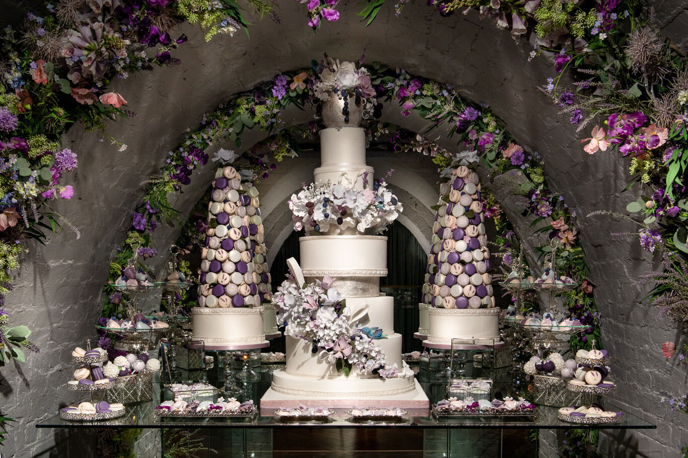 a luxury tall wedding cake surrounded by sweet deserts and purple flowers in london