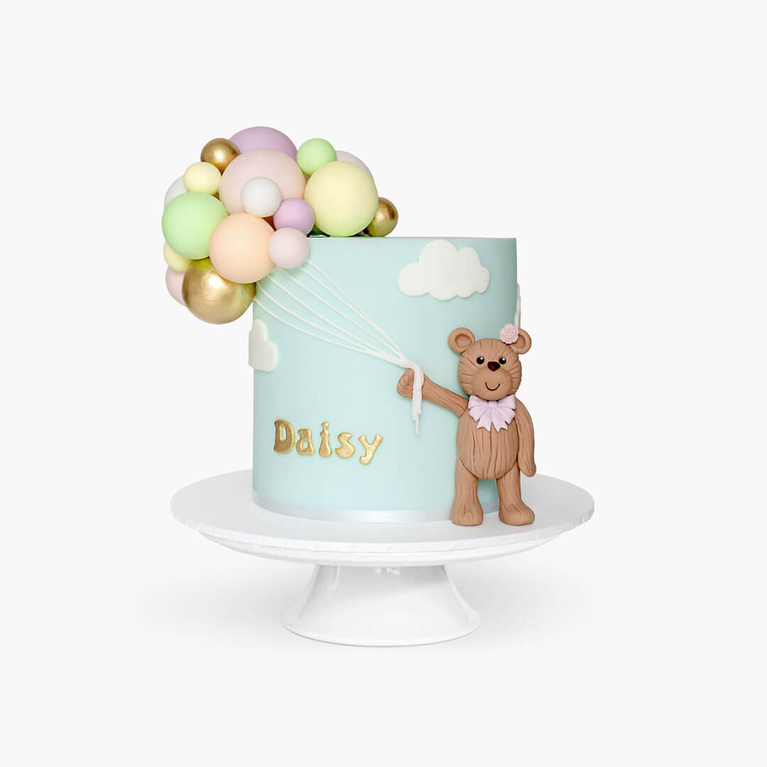 Birthday cake with a cluster of colourful balloons and a teddy bear holding onto balloon strings