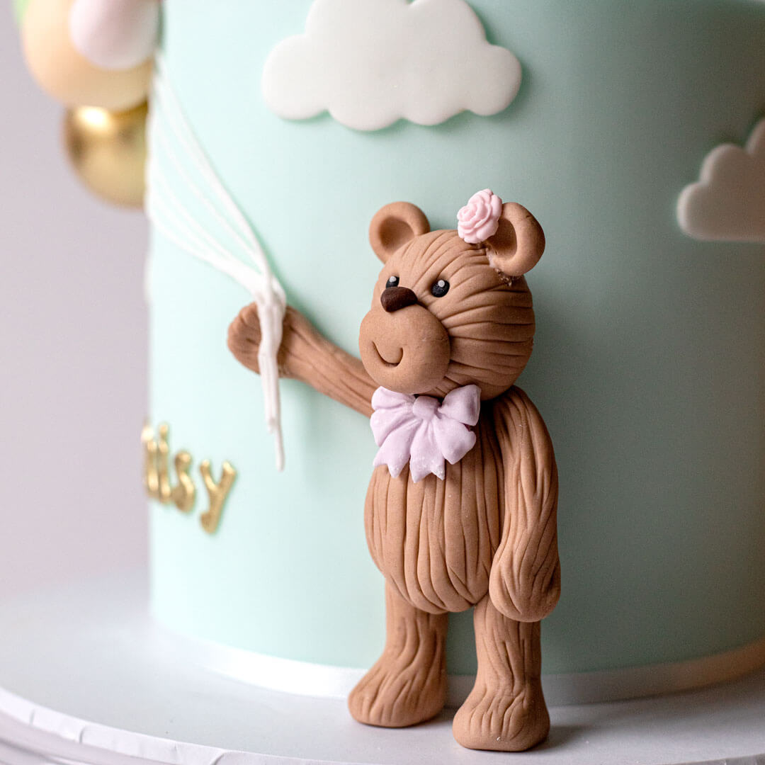 Birthday cake with a cluster of colourful balloons and a teddy bear holding onto balloon strings