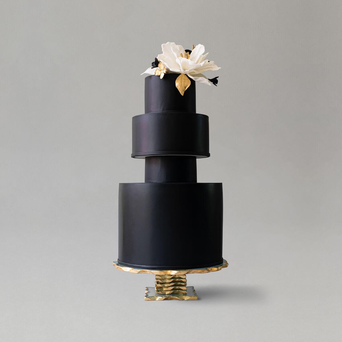 A striking black 3 tiered wedding cake topped with a large white sugar flower with gold leaf topper