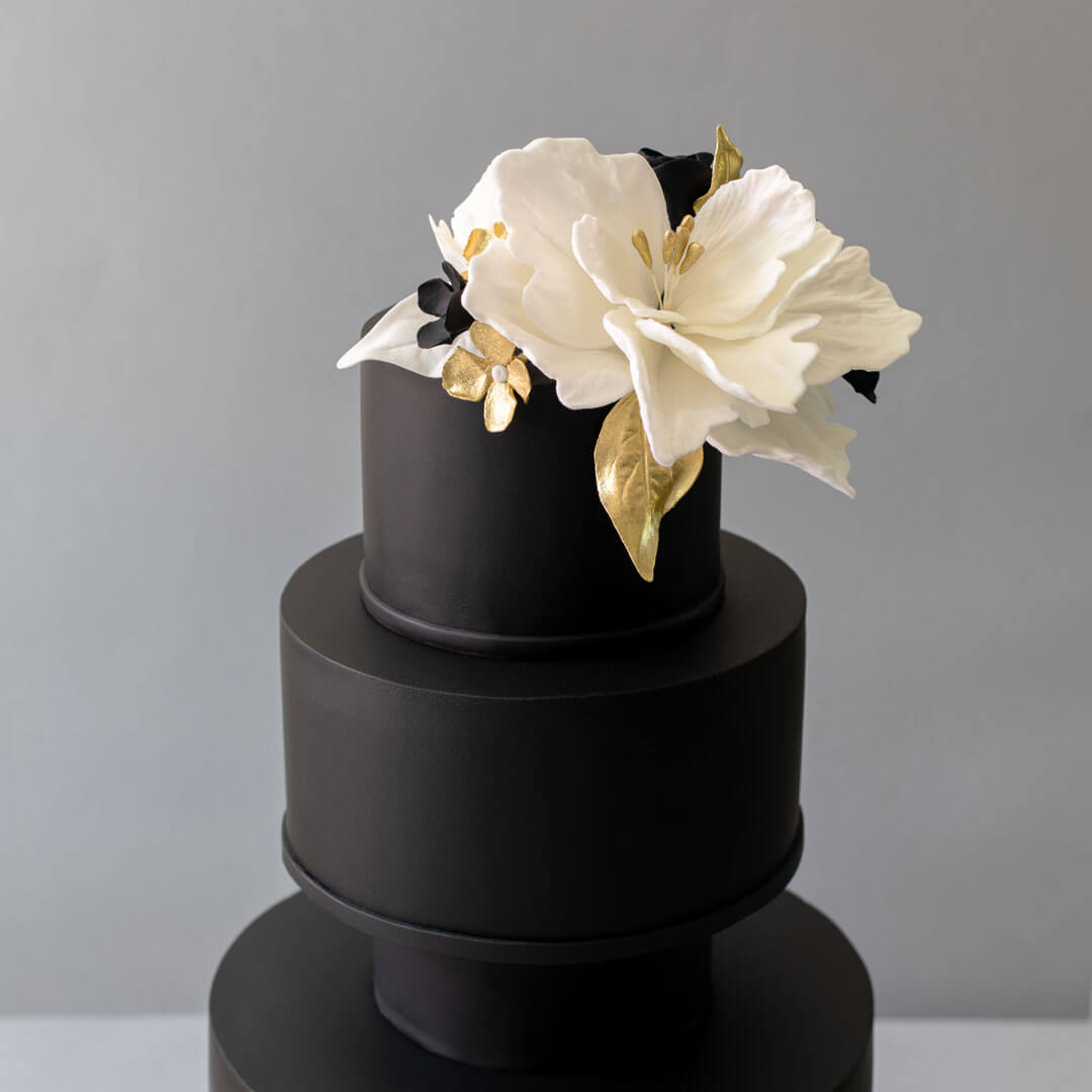 A striking black 3 tiered wedding cake topped with a large white sugar flower with gold leaf topper