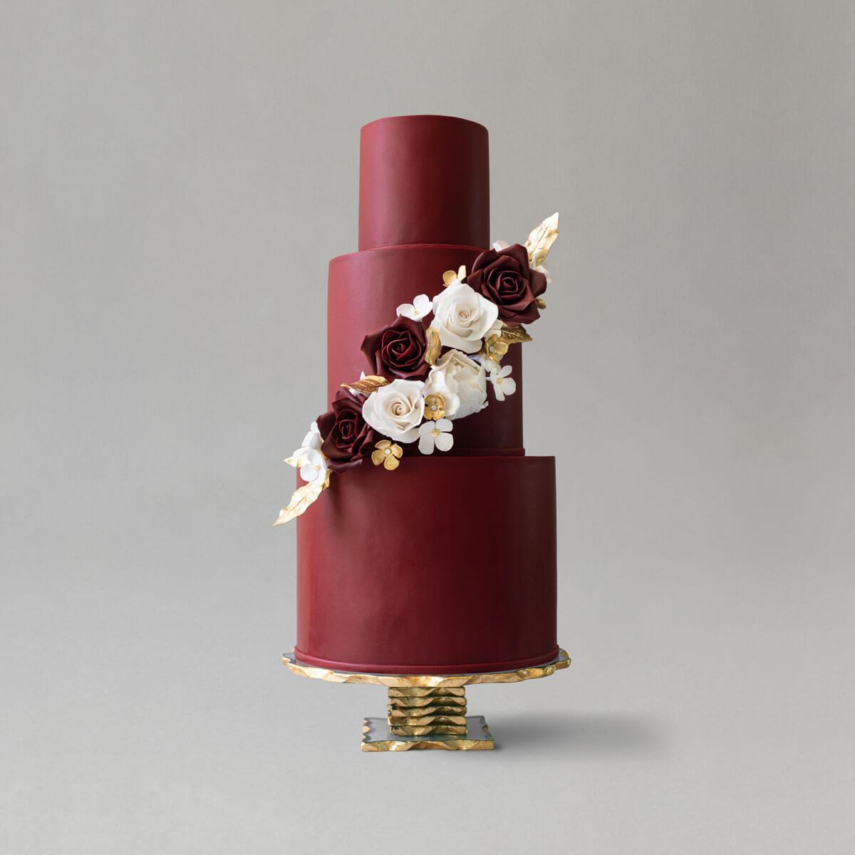 A three-tiered burgundy wedding cake decorated with cascading sugar flowers accented by gold leaves and blossoms