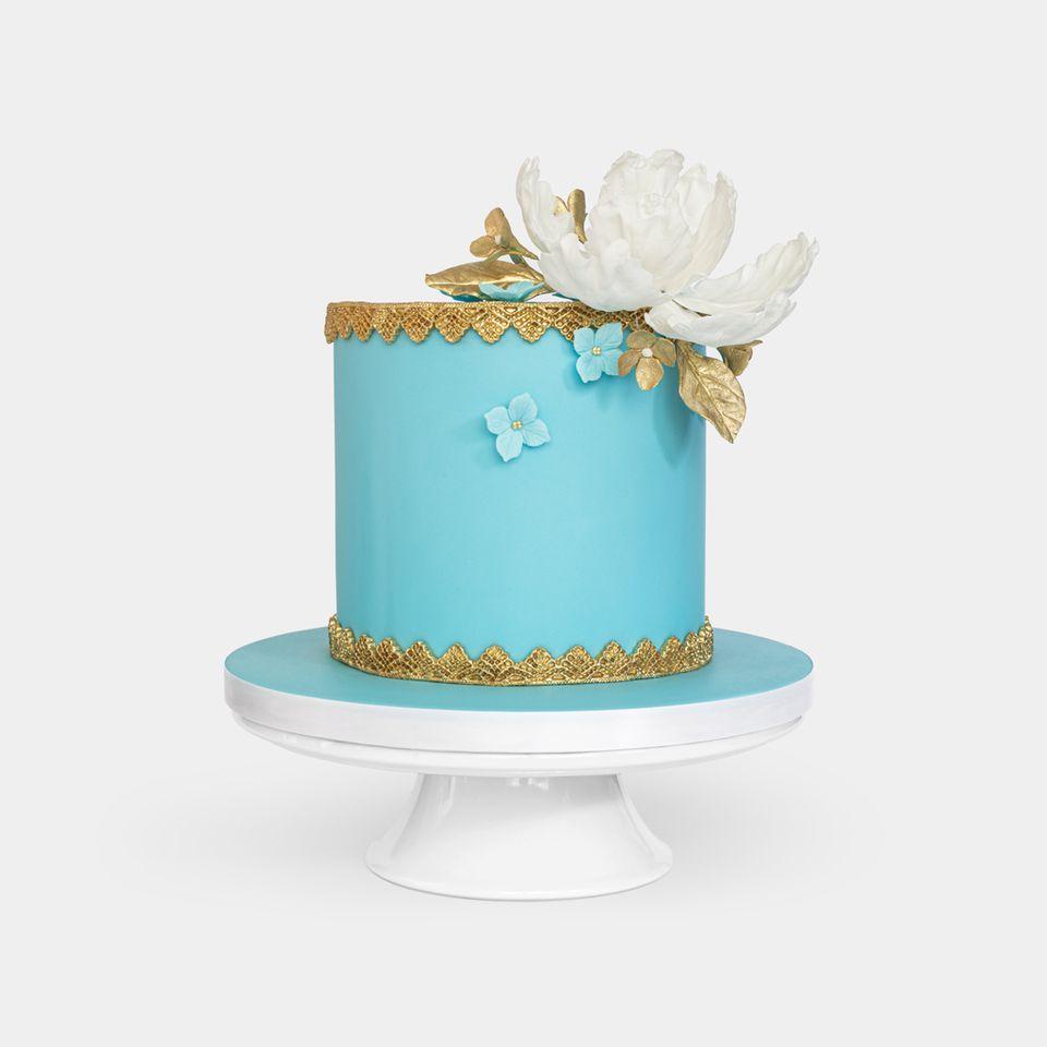 Luxurious blue fondant cake with gold lace detailing, large white peony with gold leaves topper