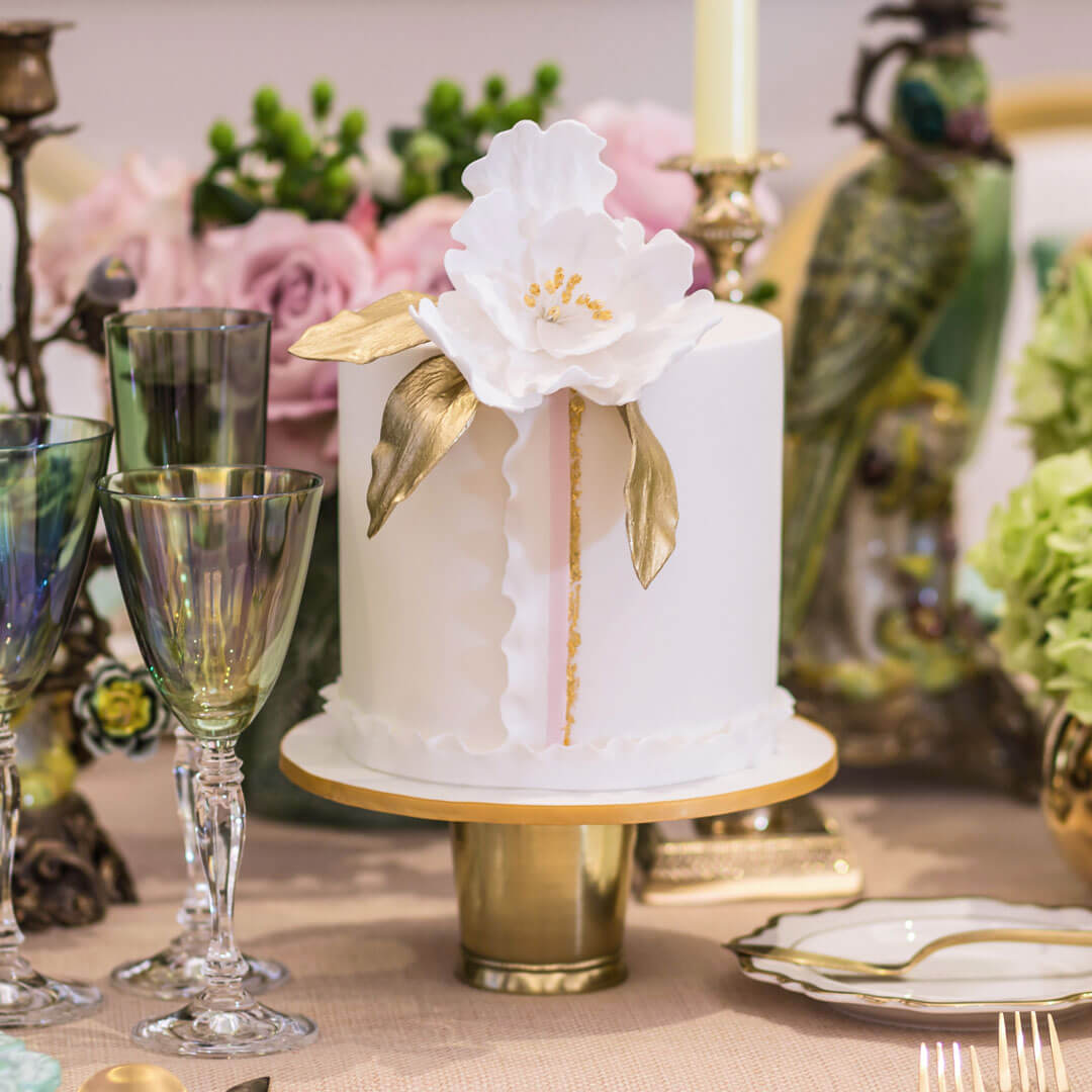 Elegant single tier white cake with a handcrafted sugar flower, gold accented leaves and delicate fondant ruffle detail