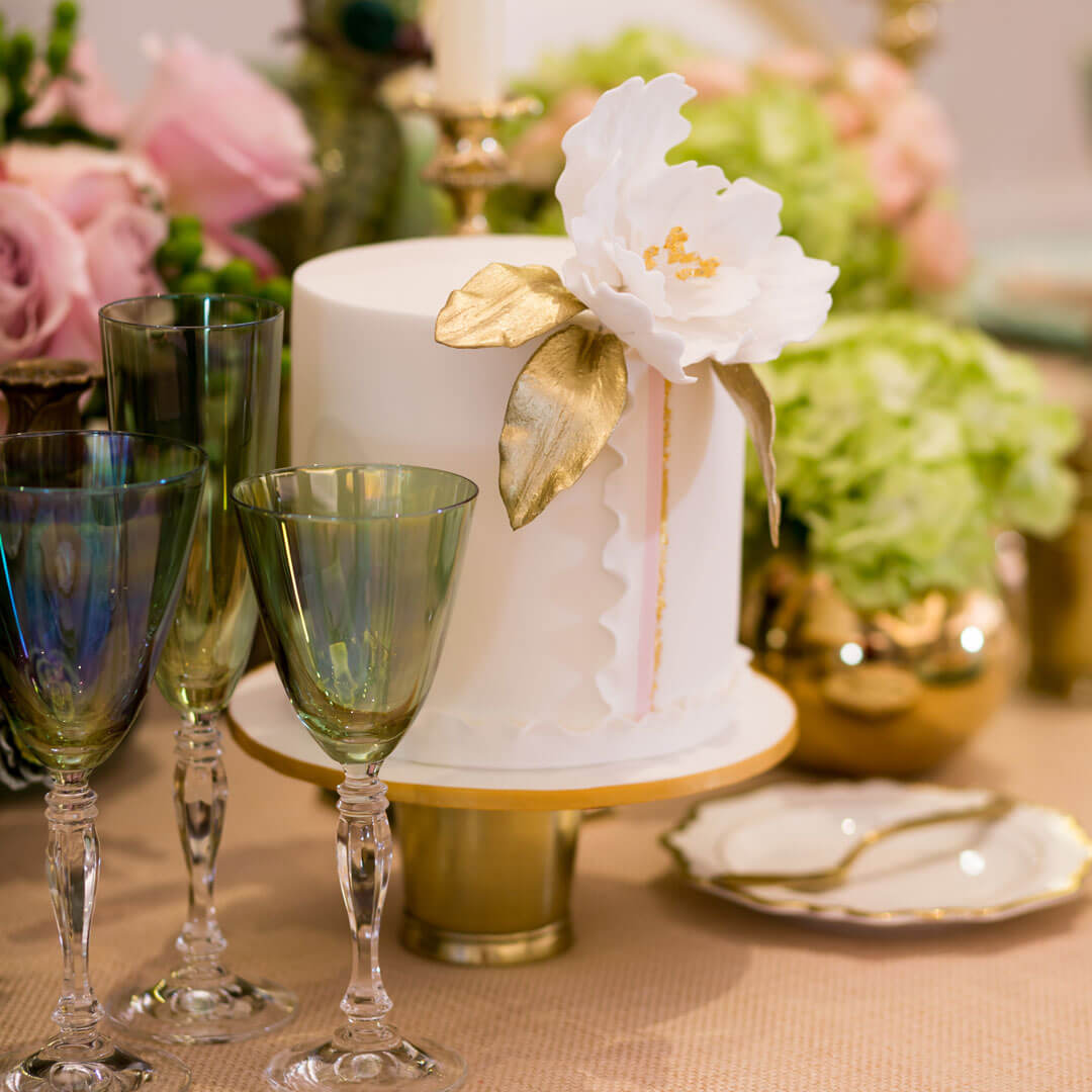 Elegant single tier white cake with a handcrafted sugar flower, gold accented leaves and delicate fondant ruffle detail