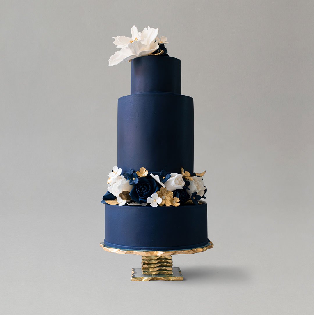 Elegant navy blue wedding cake with handmade sugar flowers between base tiers, topped with a large white handmade floral on the top