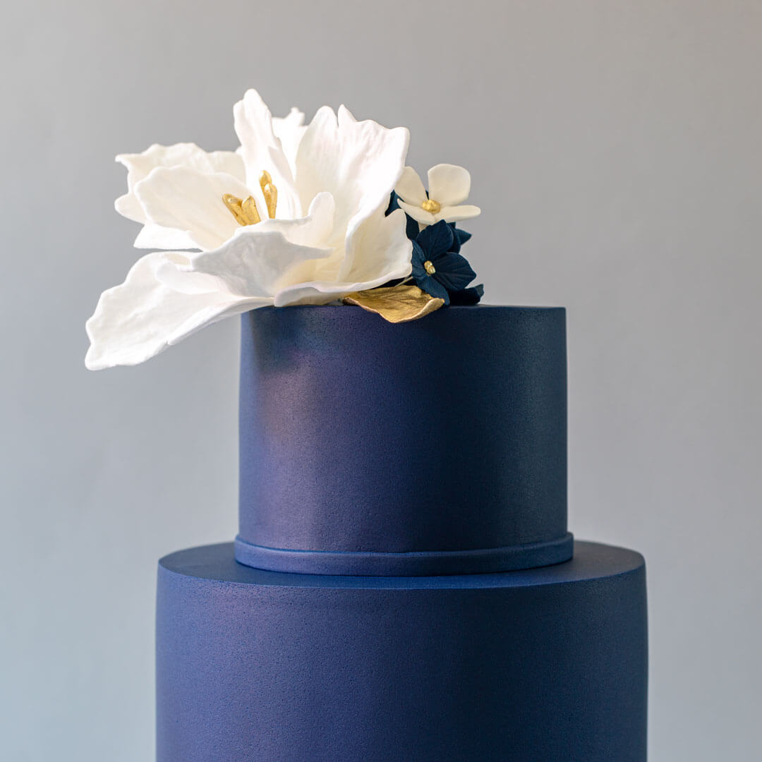 Elegant navy blue wedding cake with handmade sugar flowers between base tiers, topped with a large white handmade floral on the top